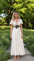 Front view of woman wearing ivory V-neck lace trim maxi dress with bow accent outdoors